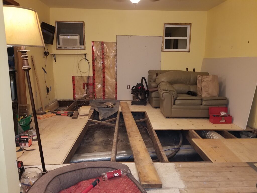 Very cluttered room with some plywood on floor but floor joists still showing